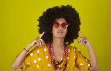 Kıvırcık saçlı, kalp şeklinde gözlüklü, sarı arka planı olan ve parmaklarıyla yönü gösteren genç ve güzel bir kadın.