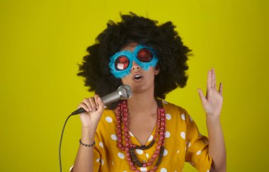 Kıvırcık saçlı, güzel ve gülümseyen bir kız ve sarı arka planda mikrofonla şarkı söyleyen komik gözlükler.