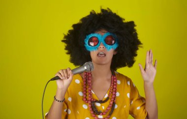 Kıvırcık saçlı, güzel ve gülümseyen bir kız ve sarı arka planda mikrofonla şarkı söyleyen komik gözlükler.