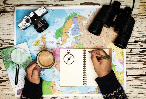 Flat lay of the travel planning concept. Top view of man hands writing on a logbook on the table binoculars, compass, retro photo camera, coffee, and Europe map on a white wooden table