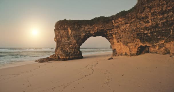 Letecký pohled na slunce nad skalní stěnou s otvorem na písečné pláži mořského pobřeží. Unikátní geologická formace — Stock video