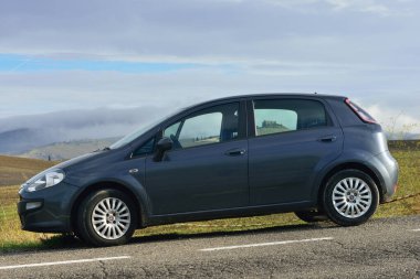 Pienza, İtalya - 13 Ekim 2012: Fiat Punto Evo arabası, sonbahar tahıl tarlaları ve araba pencerelerinden görünen eski kalesi olan tepeleri olan harika bir manzaranın önüne park etti. Toskana İtalya 'nın dünyaca ünlü Vall d' Orcia bölgesi. Seyahat noktaları konsepti