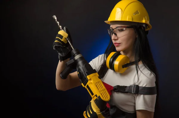 the brunette girl in special clothes and a worker in a helmet posing on a black background with a working tool ( perforator, drill, percussion hammer )