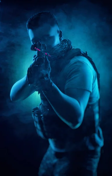 El hombre de ropas especiales militares posando con un arma en sus manos sobre un fondo oscuro en la niebla —  Fotos de Stock