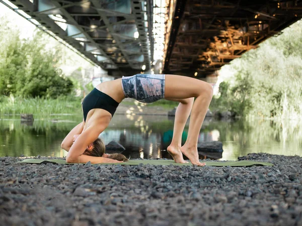 ince güzel genç kız Mat üzerinde yaz aylarında köprünün altında nehir kenarında yoga yapıyor