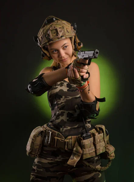 A menina em roupas militares airsoft posa com uma arma em suas mãos em um fundo escuro — Fotografia de Stock