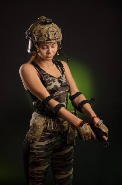 Askeri giysili kız elinde silahla karanlık bir arka planda poz veriyor.