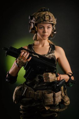 Askeri giysili kız elinde silahla karanlık bir arka planda poz veriyor.