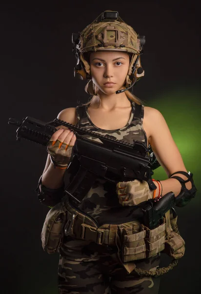 A menina em roupas militares airsoft posa com uma arma em suas mãos em um fundo escuro — Fotografia de Stock