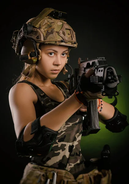 A menina em roupas militares airsoft posa com uma arma em suas mãos em um fundo escuro — Fotografia de Stock