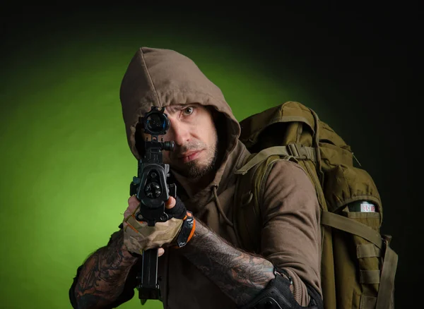 Muž stalker s pistolí s optickým zaměřovačem a batohem na tmavém pozadí — Stock fotografie