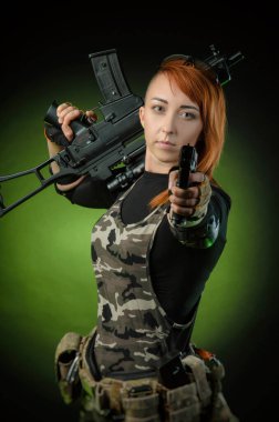 Askeri giysili kız elinde silahla karanlık bir arka planda poz veriyor.
