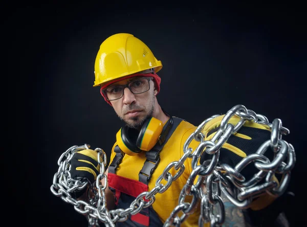 Een man in de overall van een bouwreparateur met een medisch masker tegen het coronavirus houdt een metalen ketting in zijn handen — Stockfoto