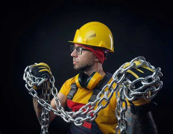 Een man in de overall van een bouwreparateur met een medisch masker tegen het coronavirus houdt een metalen ketting in zijn handen — Stockfoto