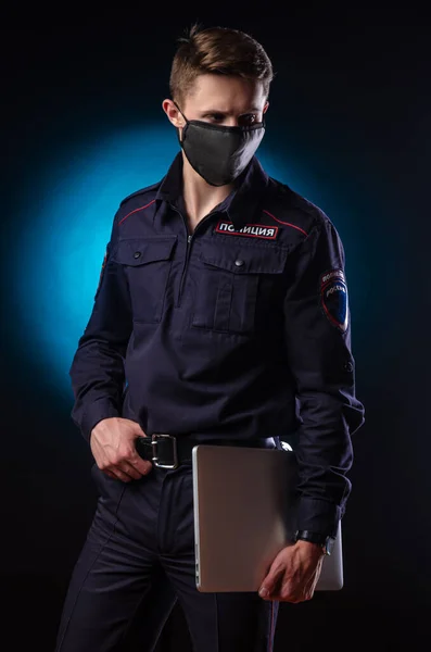 Een knappe man in een politie-uniform met een laptop op zijn gezicht en een medisch masker. Engelse vertaling van Police — Stockfoto