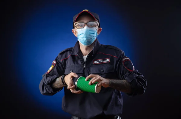 De man in het politie-uniform is een Russische politieagent met een medisch masker met een koffiebeker in zijn hand. Engelse vertaling van "Police" — Stockfoto