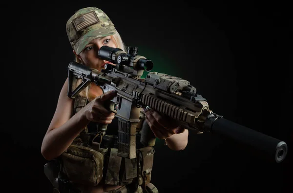 Uma menina sexy no macacão airsoft militar posa com uma arma em suas mãos em um fundo escuro — Fotografia de Stock