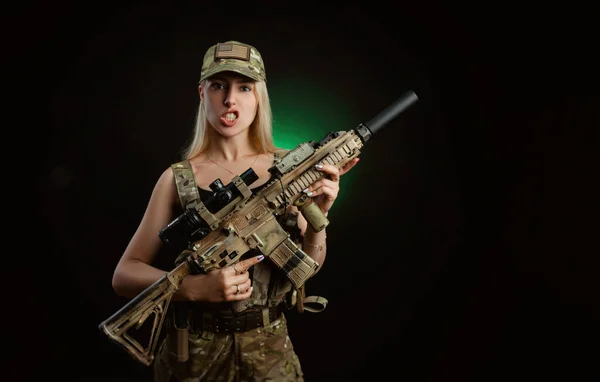 Uma menina sexy no macacão airsoft militar posa com uma arma em suas mãos em um fundo escuro — Fotografia de Stock
