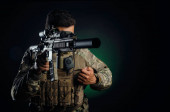 egy brutális fickó katonai airsoft overálban, fegyverrel a kezében egy sötét háttérben.