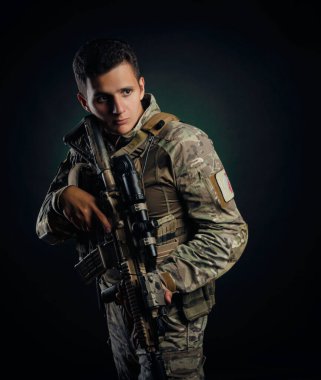 Askeri hava yumuşaklığı olan acımasız bir adam elinde silahla karanlık bir arka planda poz veriyor.