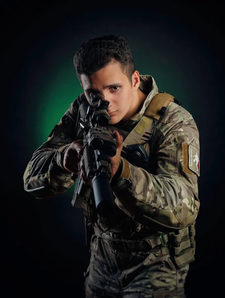Ένας κτηνώδης τύπος με στρατιωτική φόρμα ποζάρει με ένα όπλο στα χέρια του σε σκοτεινό φόντο. — Φωτογραφία Αρχείου