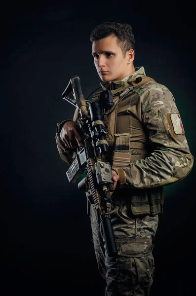 Ένας κτηνώδης τύπος με στρατιωτική φόρμα ποζάρει με ένα όπλο στα χέρια του σε σκοτεινό φόντο. — Φωτογραφία Αρχείου
