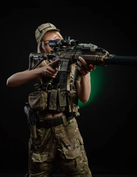 Uma menina sexy no macacão airsoft militar posa com uma arma em suas mãos em um fundo escuro — Fotografia de Stock