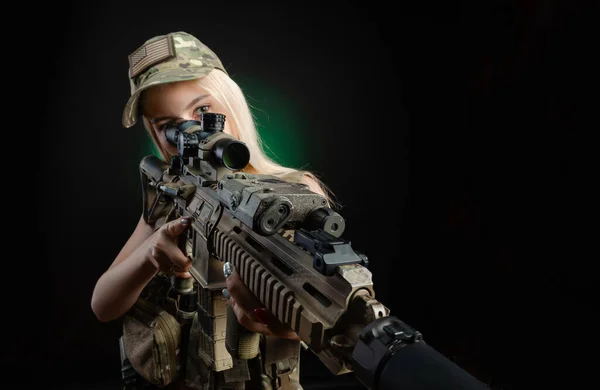 Uma menina sexy no macacão airsoft militar posa com uma arma em suas mãos em um fundo escuro — Fotografia de Stock