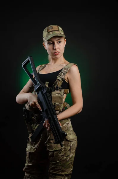Una chica sexy en overoles militares de airsoft posa con un rifle de asalto sobre un fondo oscuro —  Fotos de Stock