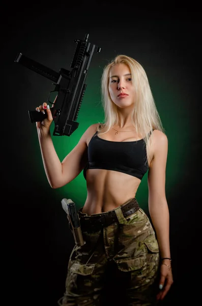 Una chica sexy en overoles militares de airsoft posa con un rifle de asalto sobre un fondo oscuro — Foto de Stock