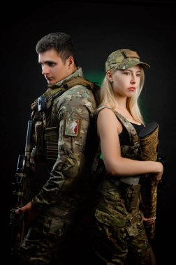 Askeri tulumlu bir kız ve bir adam koyu bir arka planda hava yumuşaklığında bir silahla poz veriyorlar.