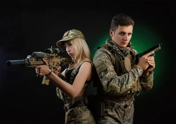 Uma menina e um cara em macacão militar posar com uma arma airsoft em um fundo escuro — Fotografia de Stock