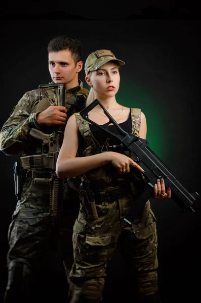 Uma menina e um cara em macacão militar posar com uma arma airsoft em um fundo escuro — Fotografia de Stock