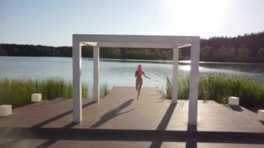 Mayolu bir kız iskelede koşuyor. Güneş parlıyor.