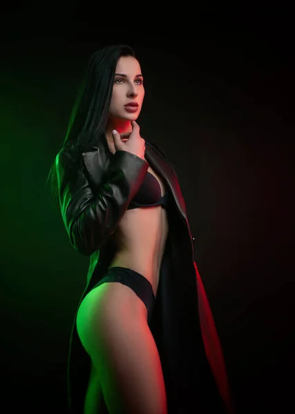 Beautiful hot brunette girl posing in a photo Studio on a dark background d leather raincoat neon light — Stock Photo, Image