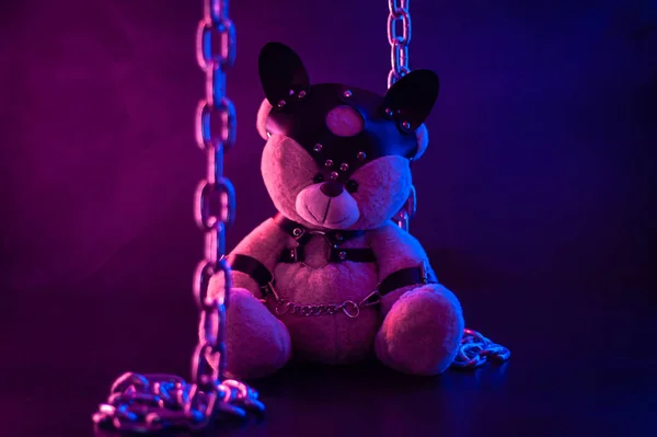 toy bear dressed in leather belts harness accessory for BDSM games on a dark background in neon light