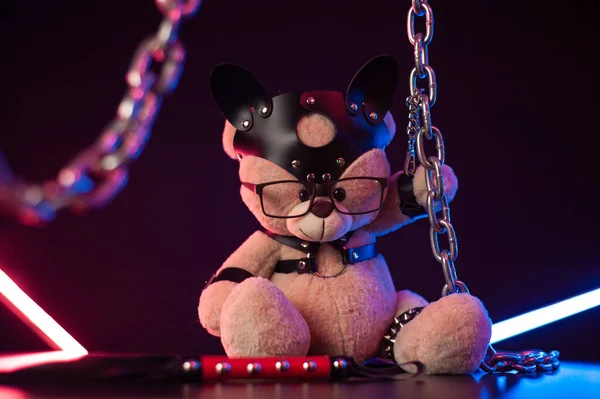 Toy bear dressed in leather belts harness accessory for BDSM games on a dark background in neon light — Stock Photo, Image