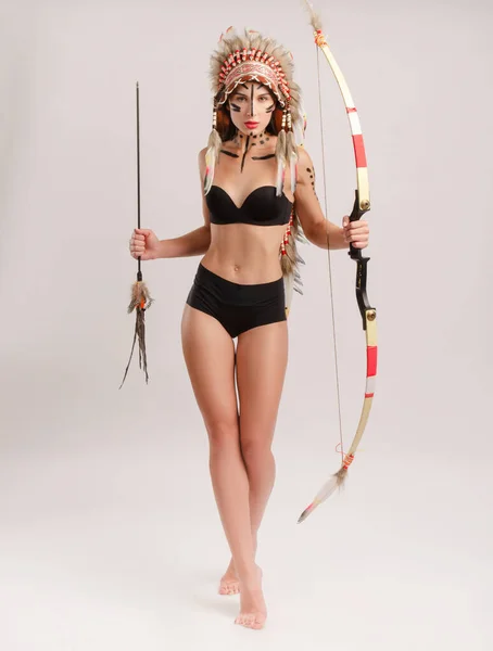 Girl in the image of indigenous peoples of America with a bow and arrow posing on a light background — Stock Photo, Image