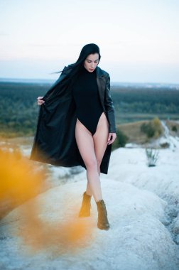 Siyah bir beden ve deri yağmurluk giymiş ince bir esmer doğada cinsel bir poz veriyor.