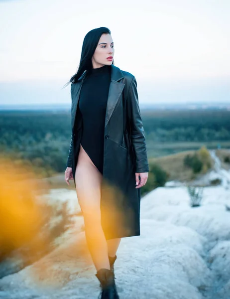A slender brunette in a black bodysuit and leather raincoat poses sexually in nature — Stock Photo, Image