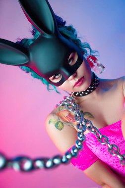 Parlak pembe elbiseli ve tavşan maskeli bir kadın parlak bir arkaplanda poz veriyor.