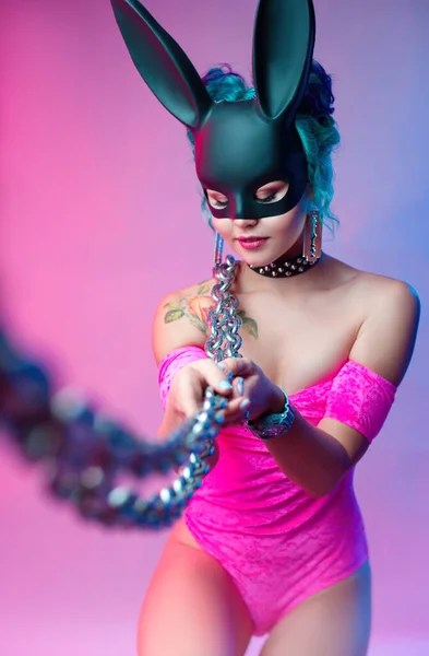 Una mujer en un traje de cuerpo de color rosa brillante y máscara de conejo posa sobre un fondo brillante con una cadena —  Fotos de Stock