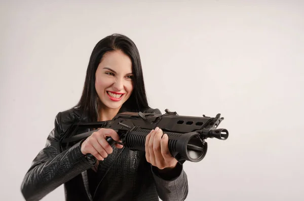 Una mujer morena posa en una chaqueta de cuero con la espalda sobre un fondo blanco con una pistola — Foto de Stock