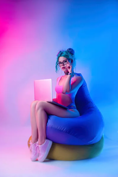 Kobieta w jasnym stroju kąpielowym z niebieskimi włosami siedzi na torbie na krześle z laptopem — Zdjęcie stockowe