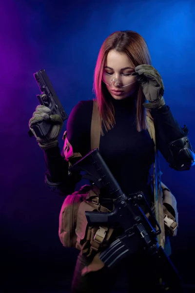 Una mujer en uniforme militar de airsoft con un rifle automático americano y una pistola sobre un fondo oscuro — Foto de Stock