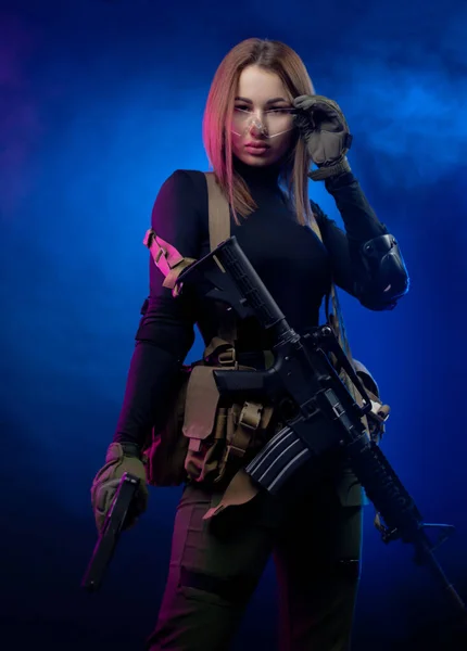 A woman in military airsoft uniform with an American automatic rifle and pistol on a dark background — Stock Photo, Image