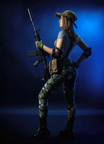 A woman in a military airsoft uniform in a vest with an American automatic rifle on a dark background — Stock Photo, Image