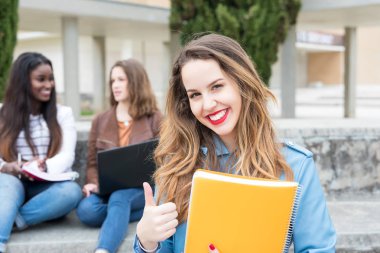 Gülümseyen öğrenci üniversite kampüsü içinde başparmak gösterilmesini