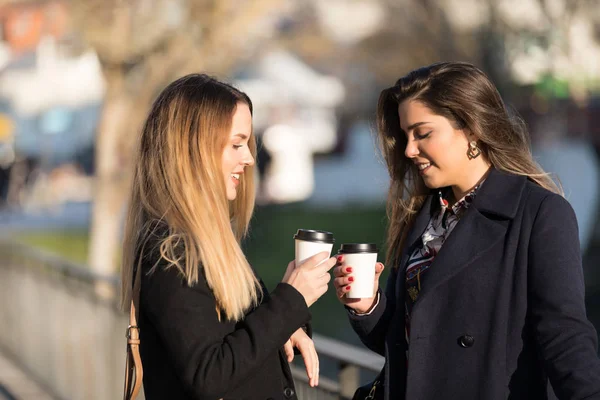 Outdoors fashion portrait of two young beautiful women friends drinking coffee. Smiling and going shopping. Kissing a cup of coffee. Bright make up