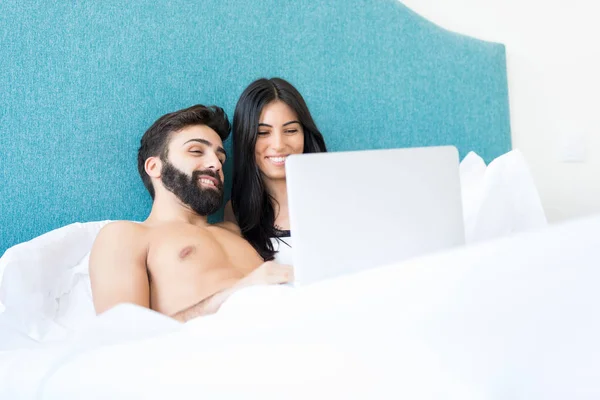 Happy couple watching on laptop screen while relaxing in bed